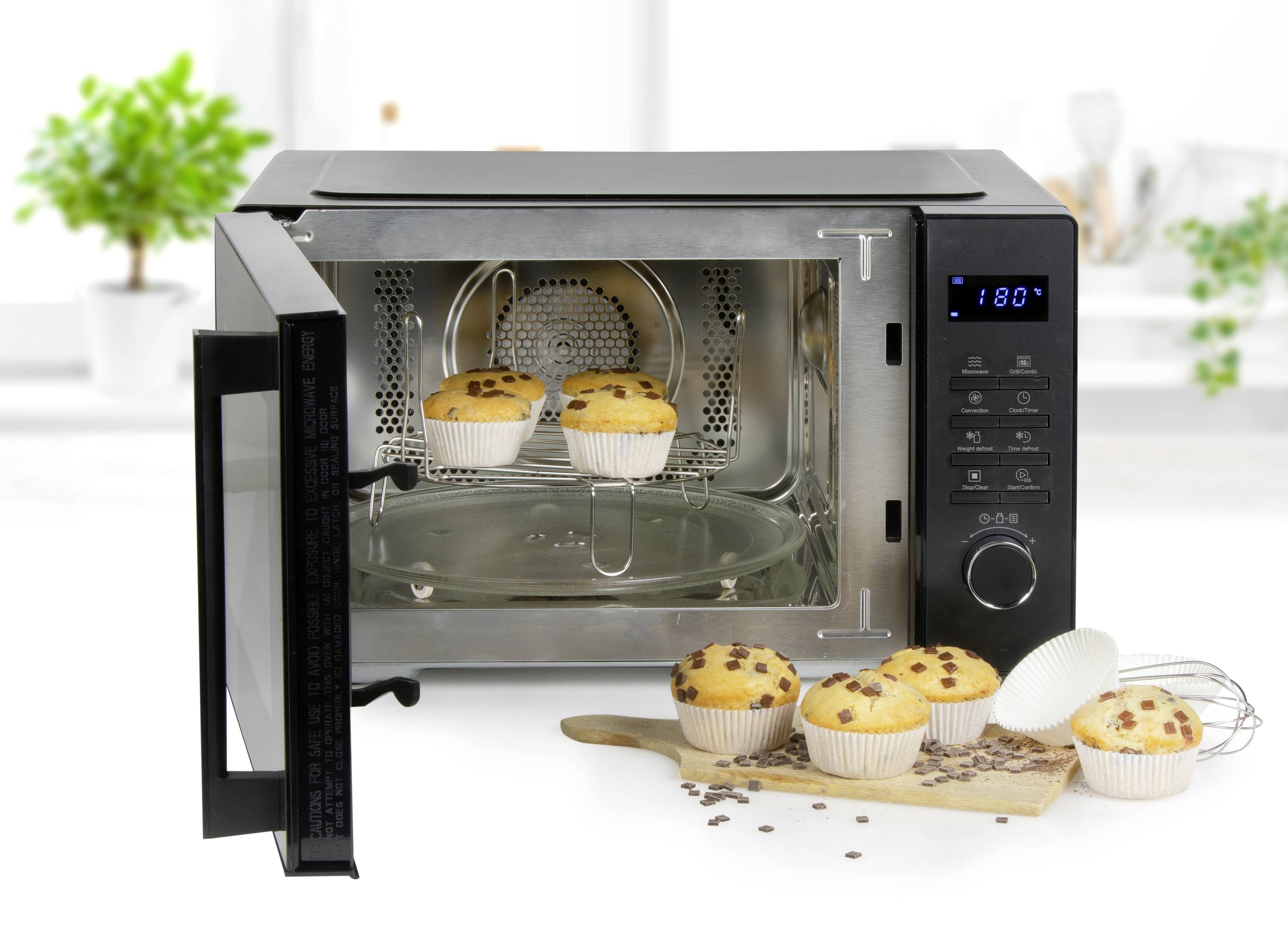 A microwave with an open door shows three muffins inside, with additional muffins placed next to it on a chopping board.