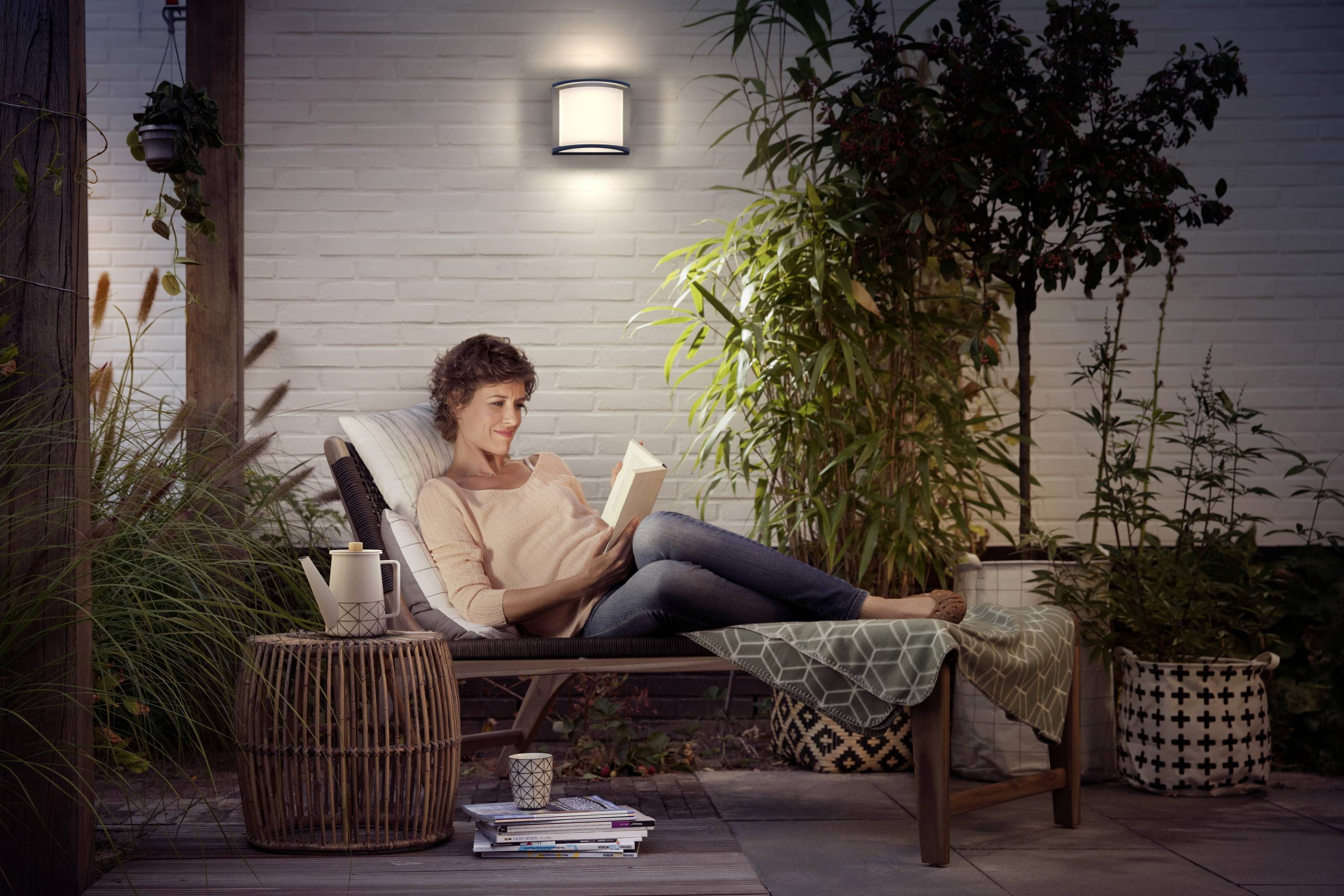 A woman is lying on a lounger in a garden, reading a book. She is surrounded by plants, with evening lighting creating a soft ambience.