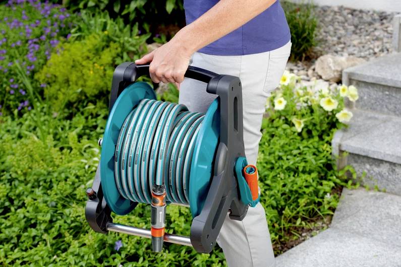 A person carries a coiled garden hose in a portable reel, walking next to greenery and stone steps.