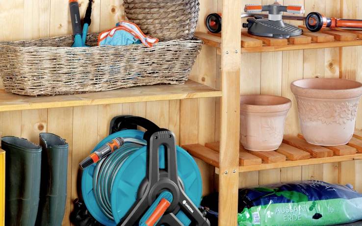 Garden shed with gardening tools, coiled hose, and empty flower pots on wooden shelves. Rubber boots stand at the bottom.