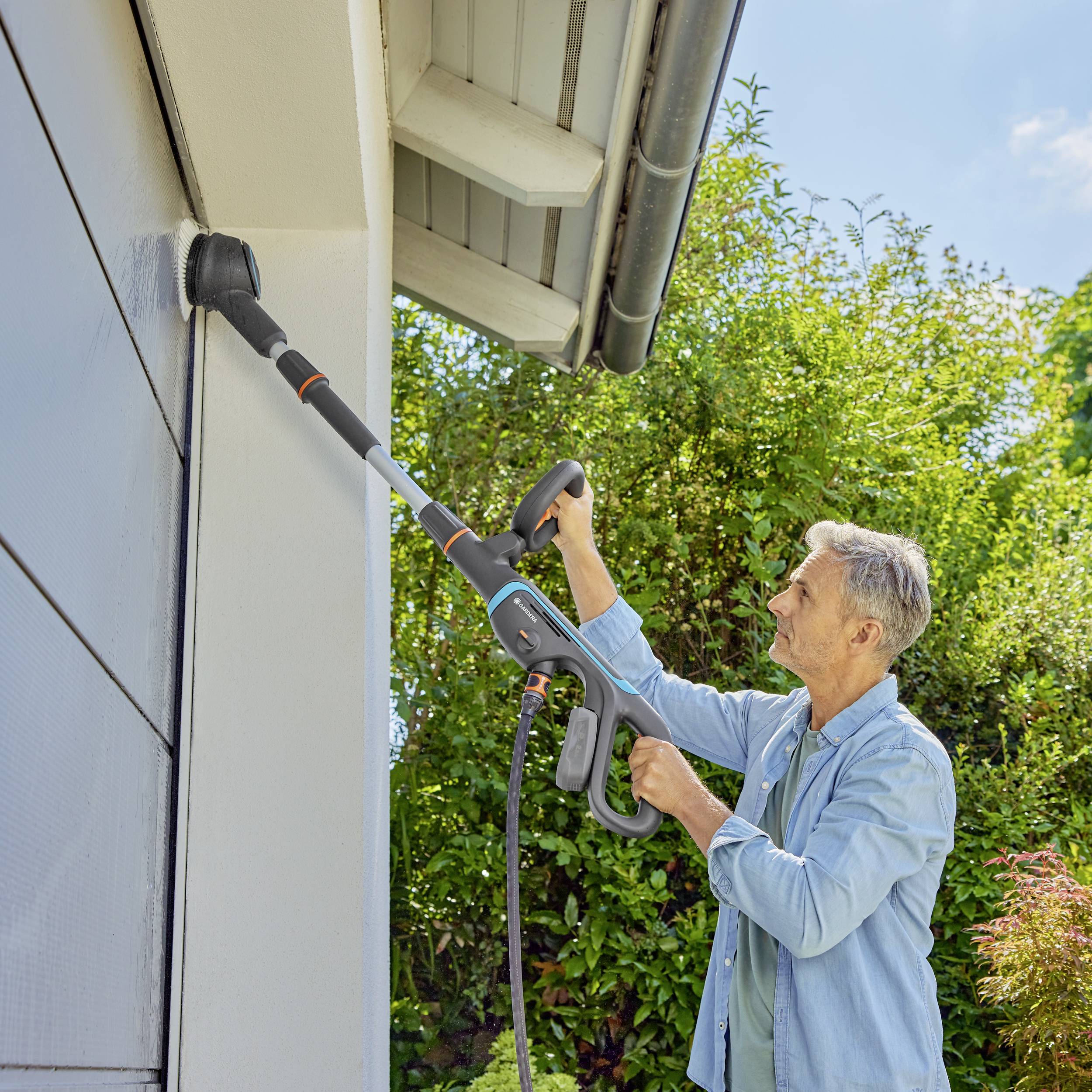 A man is cleaning the roof of a house with a portable pressure washer. Green trees in the background.
