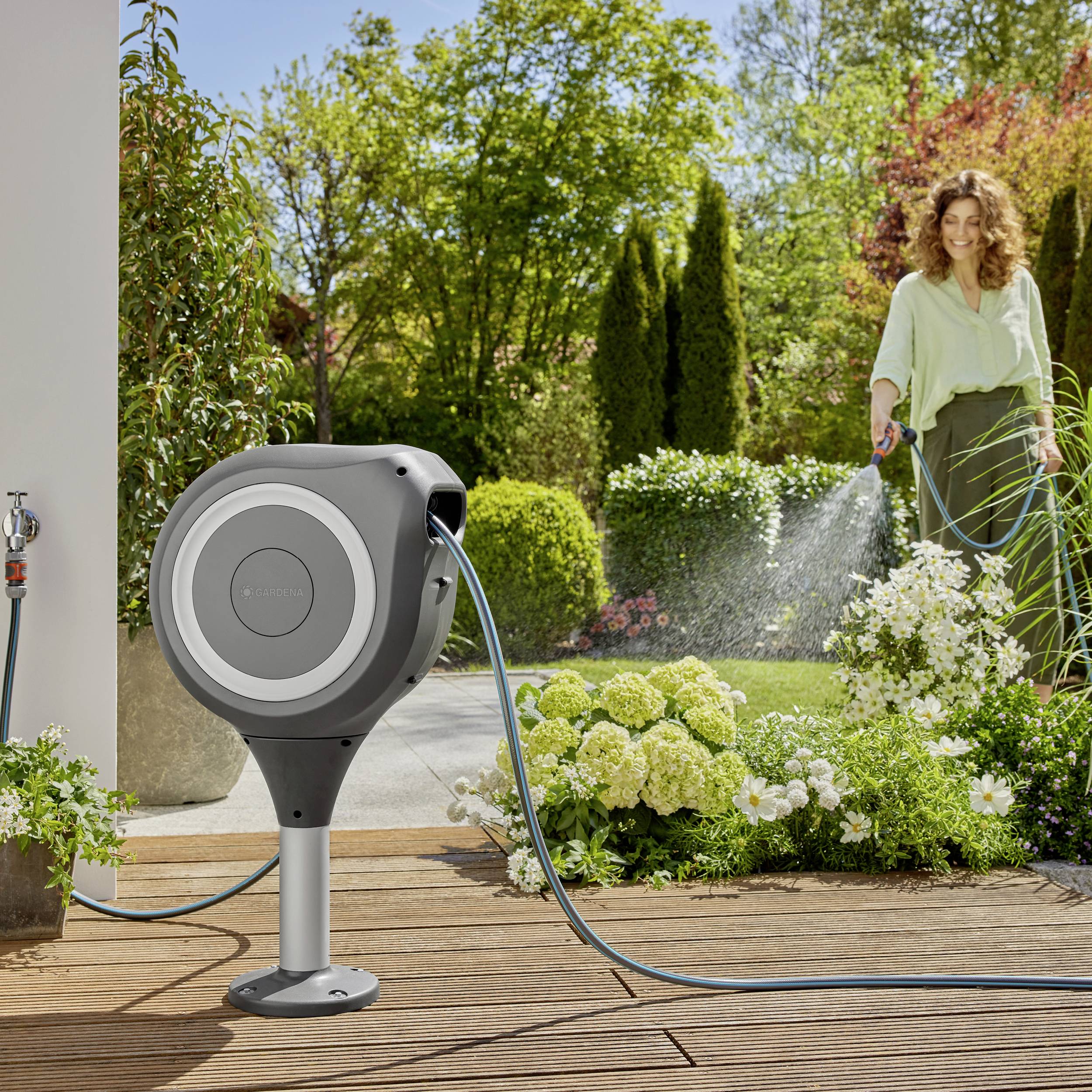 A woman is watering plants in a garden with a hose from a modern, round garden sprayer. Bright day, lots of greenery.