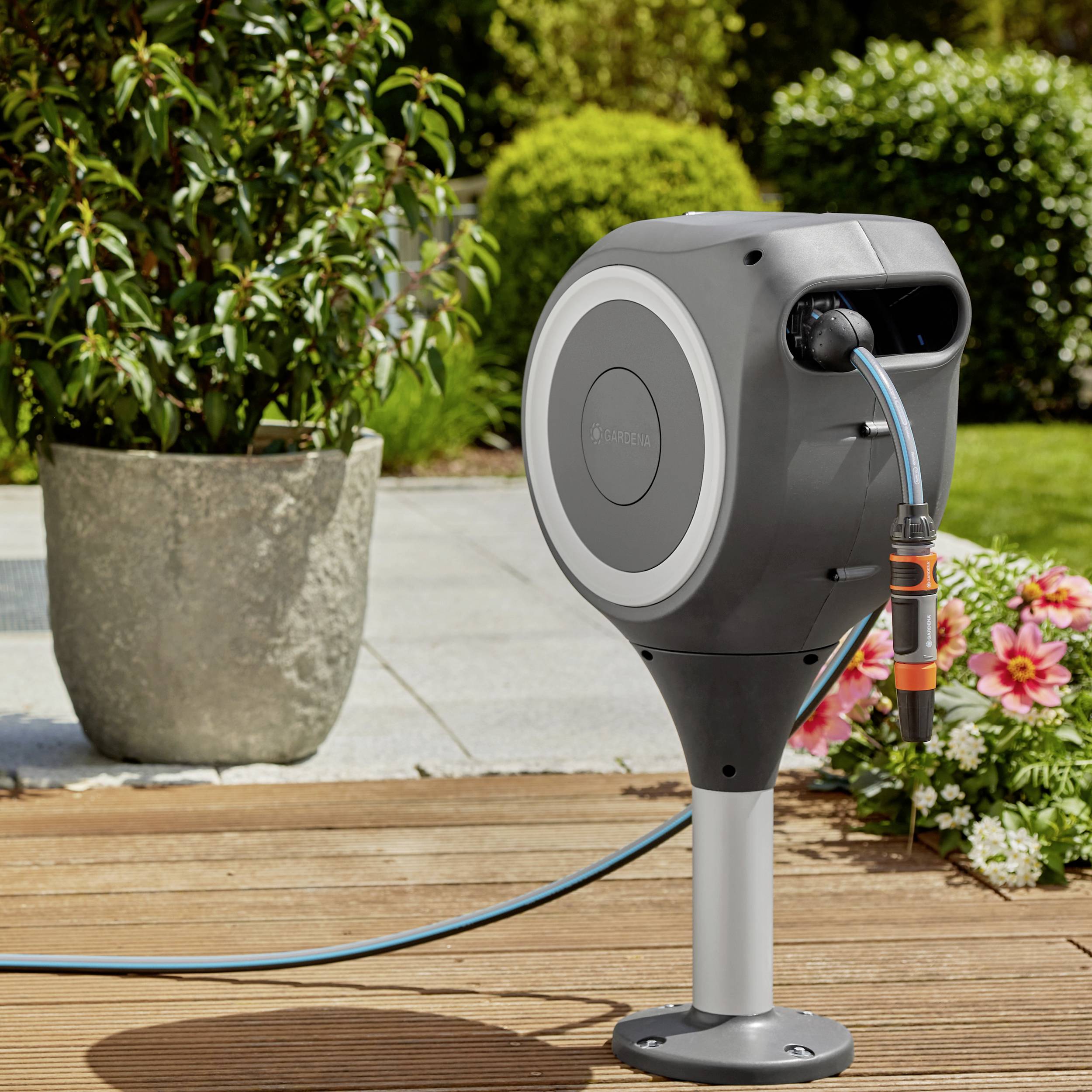 A grey garden hose reel with automatic retraction mechanism stands on a patio next to flowers and a large planter.