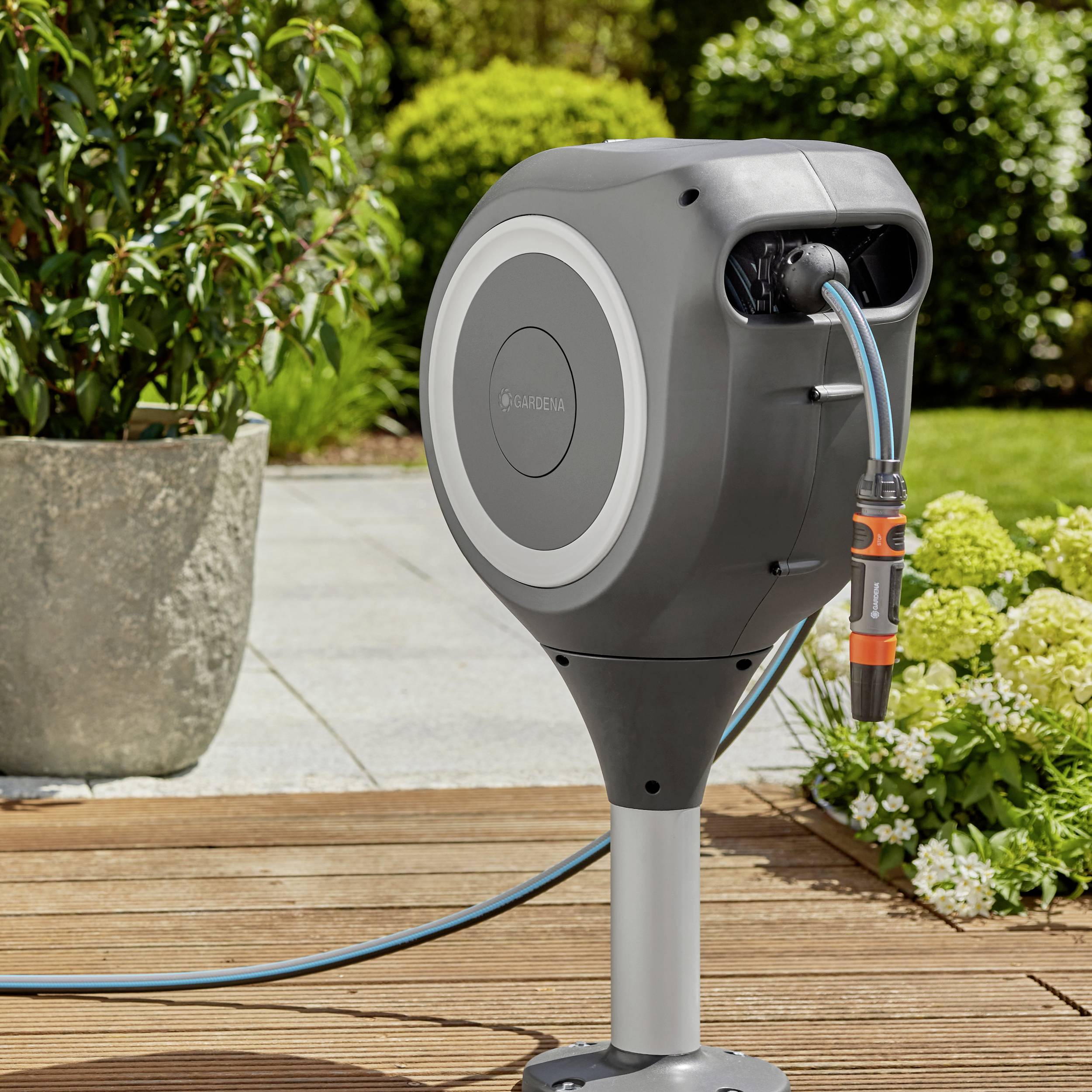 An automatic garden hose on a bracket stands in the garden. Plants and paving stones are visible in the background.