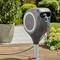 An automatic garden hose on a bracket stands in the garden. Plants and paving stones are visible in the background.