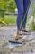 A person in jeans is cleaning the pavement with a brush and water. Garden plants are visible in the background.