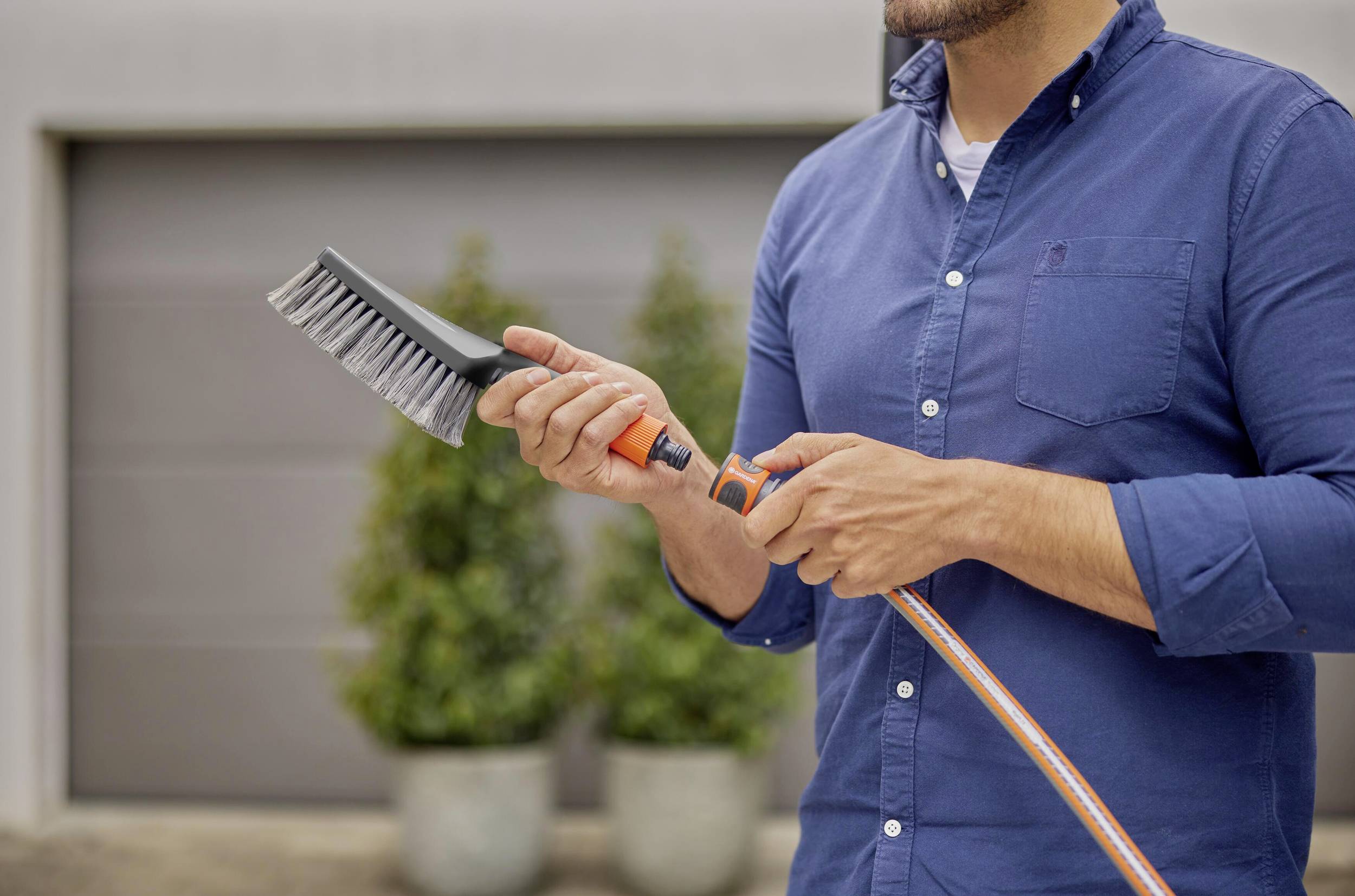 A person is holding a broom and a garden hose outdoors. Plants and a building can be seen in the background.