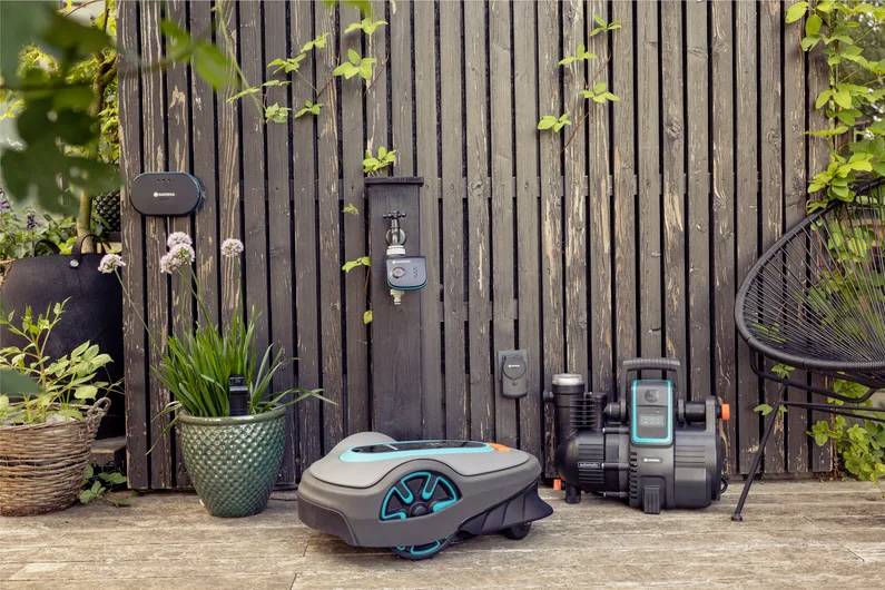 Robotic lawn mower resting on a patio near garden plants and a wooden fence, showcasing automated gardening technology in a serene setting.