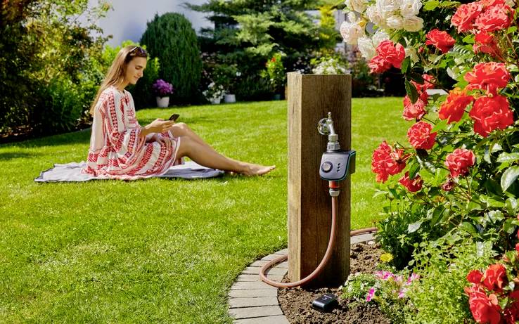 A woman in a red and white dress sits on the grass using a smartphone. A garden hose timer is attached to a tap near red and white roses.