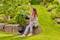 A woman sits on a stone wall in a lush garden, watering plants with a blue hose. She looks content, surrounded by greenery and flowers.