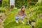 A woman crouches on a garden path using a soil moisture sensor among flowers and shrubs, checking plants' hydration levels.
