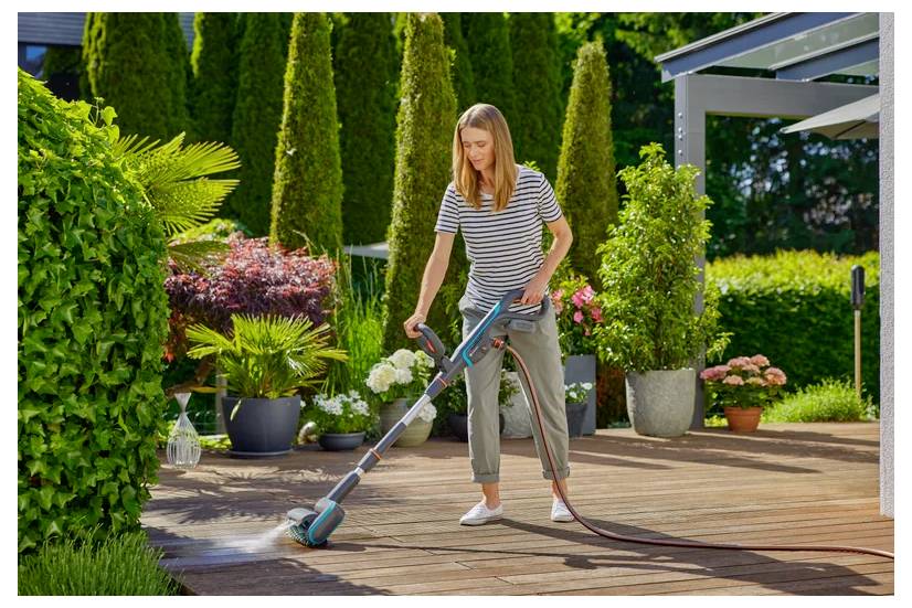 A woman uses a power washer to clean a wooden deck. Lush greenery and colorful potted plants surround her, creating a serene outdoor setting.