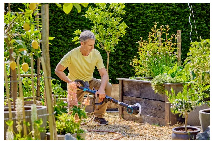 A man in a yellow shirt uses a garden sprayer to water plants and raised wooden garden boxes in a lush, well-maintained garden.