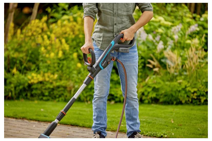A person in jeans and a long-sleeved shirt uses an electric trimmer on a neatly edged lawn, with a garden in the background.