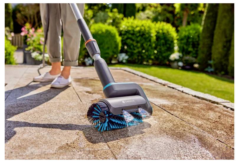 A person cleans a patio with a scrubbing tool on a sunny day, surrounded by a garden with green shrubs and flowers.