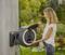 A woman uses a round wall-mounted hose reel outdoors, surrounded by flowering plants and trees.