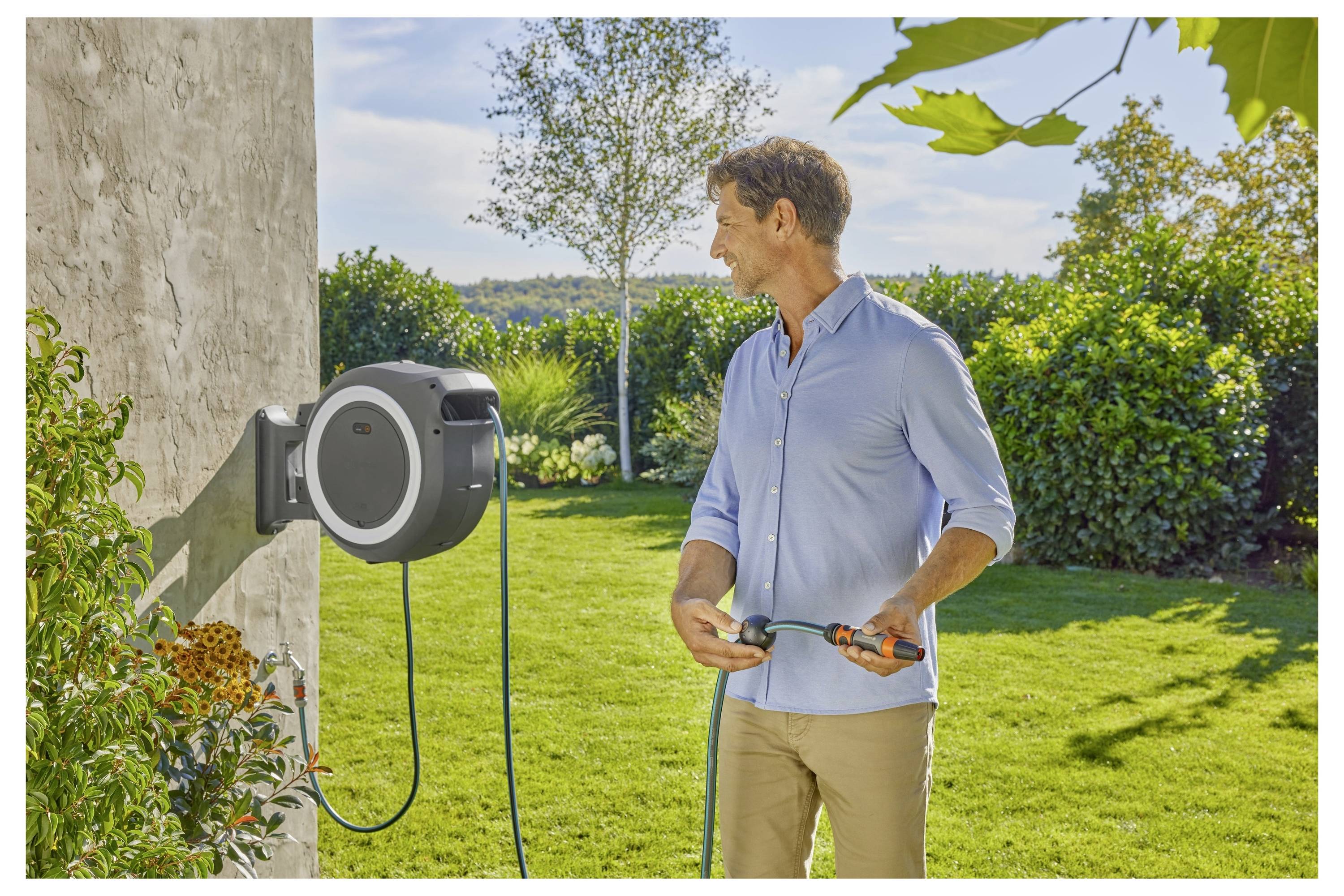 A person wearing a light blue shirt stands in a sunny garden holding a garden hose by a wall-mounted hose reel, surrounded by green shrubs.