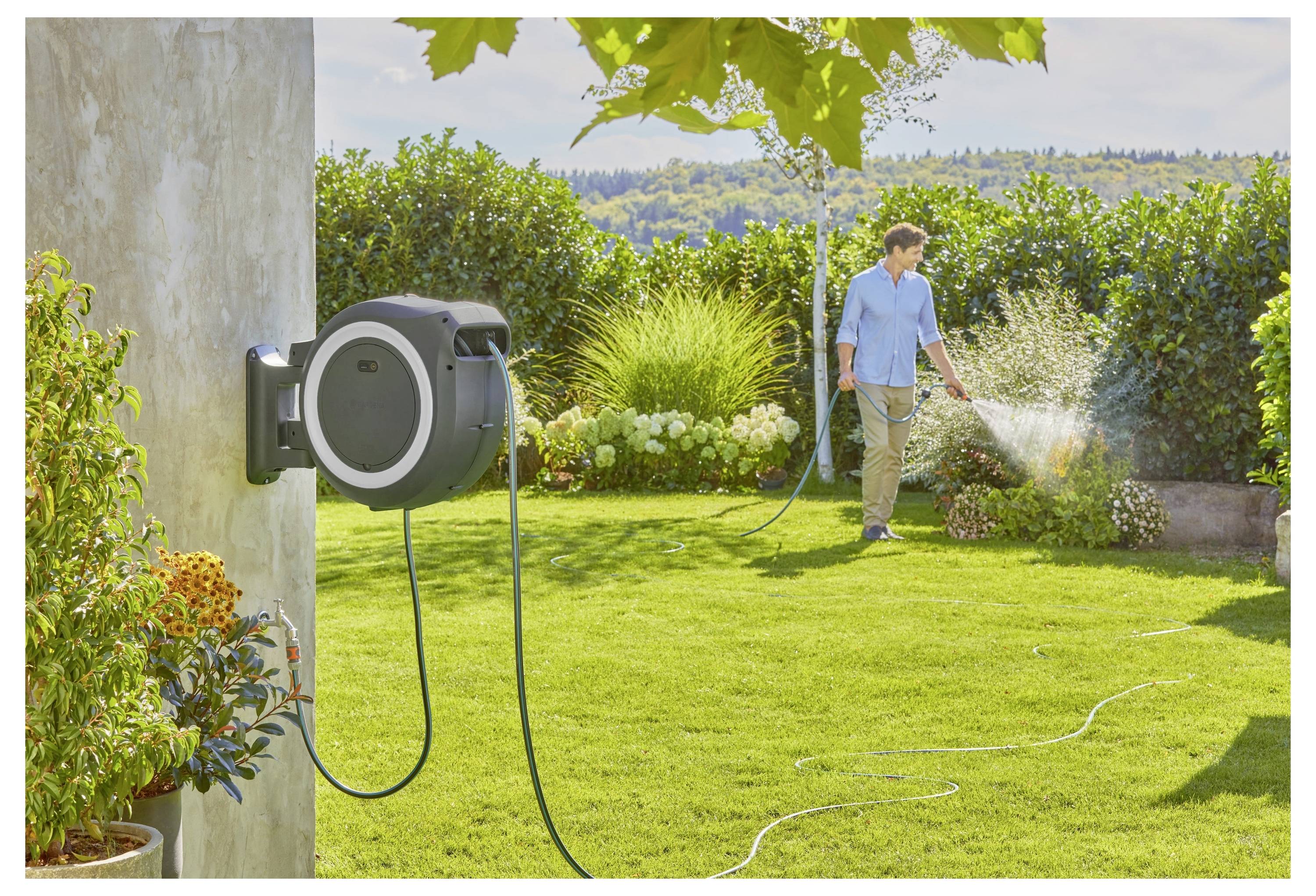 A person waters a garden with a hose attached to a wall-mounted reel. Lush greenery and blooming plants surround the yard.