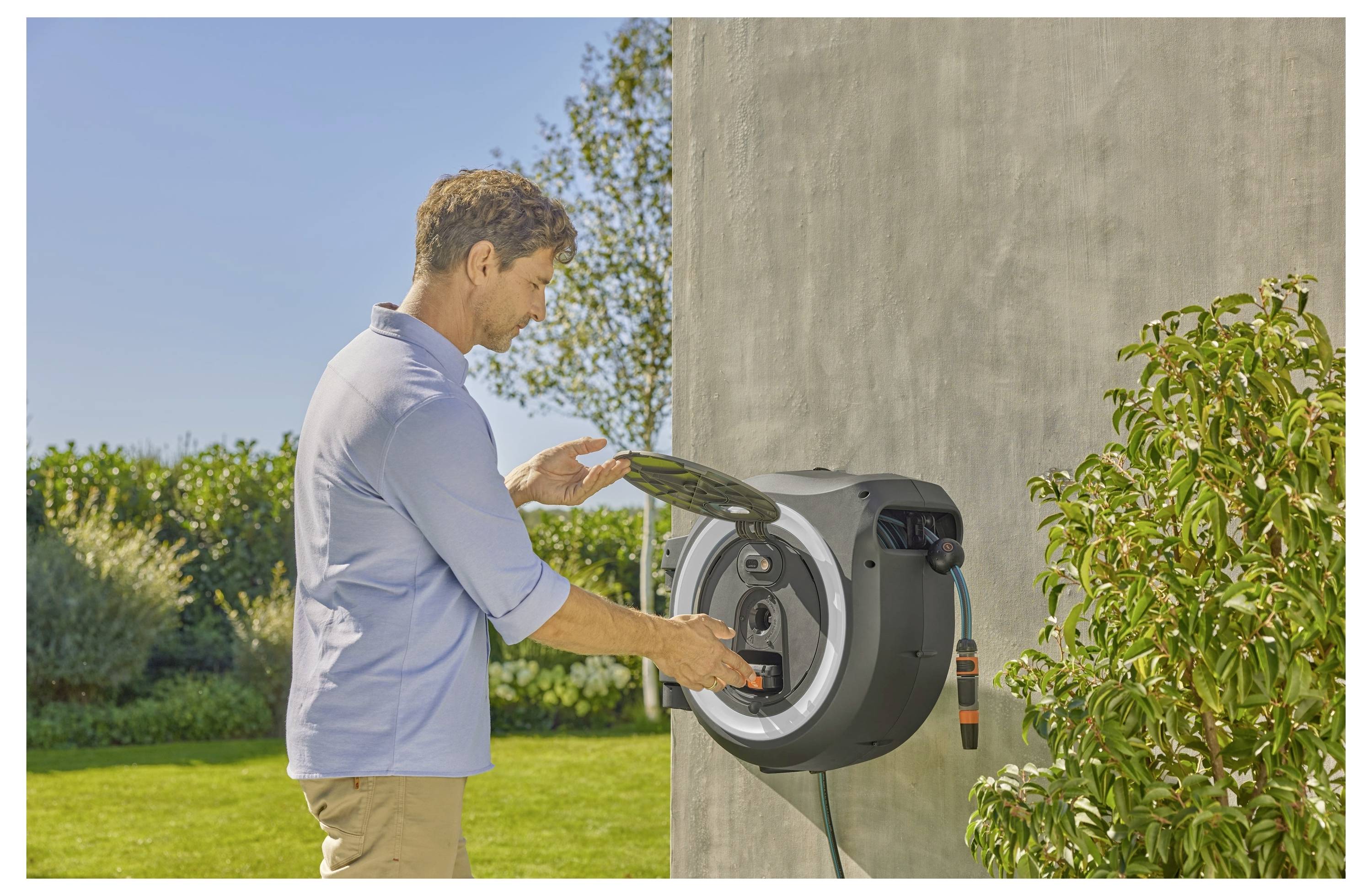 A person is operating a wall-mounted hose reel on a sunny day in a garden setting, surrounded by green plants and a clear blue sky.
