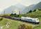 A goods train travels through a mountainous landscape. Green mountains and a cloudy sky can be seen in the background.