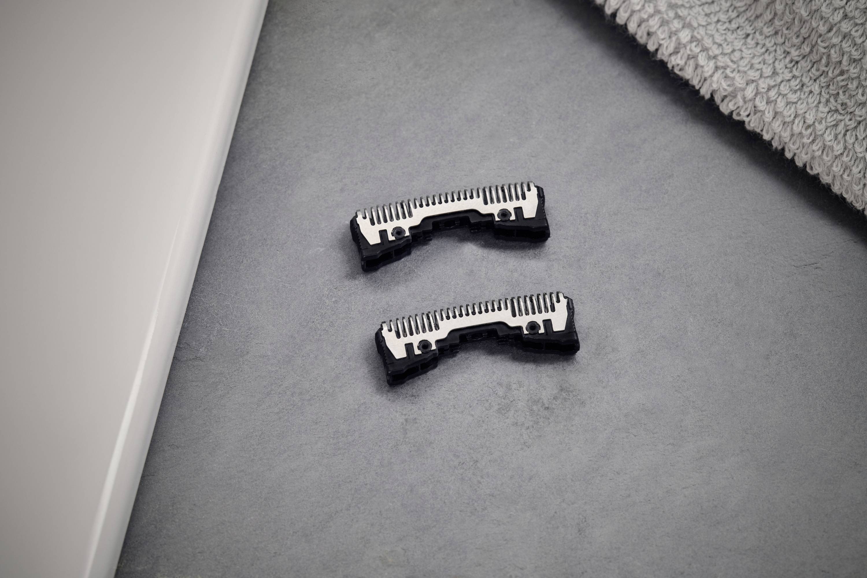 Two black and white razor heads are lying side by side on a grey surface next to a towel.