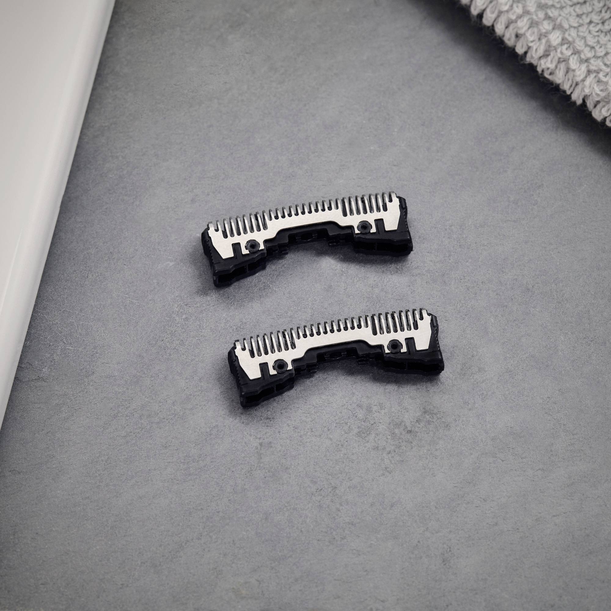 Two black and silver electric razors lie on a grey surface next to a towel.