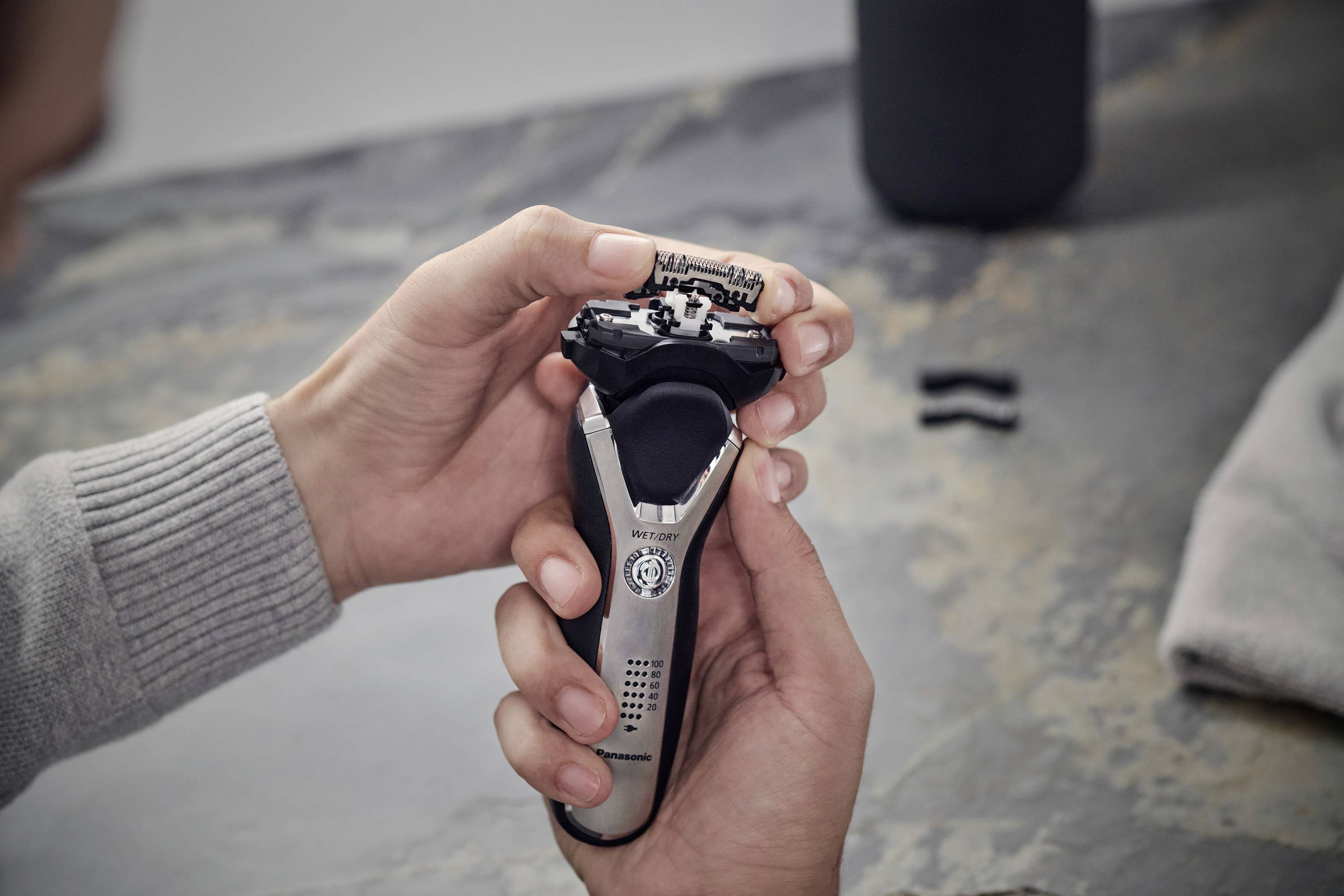 A person is holding an electric razor in their hand. The razor is silver with black accents. Blurred objects are visible in the background.