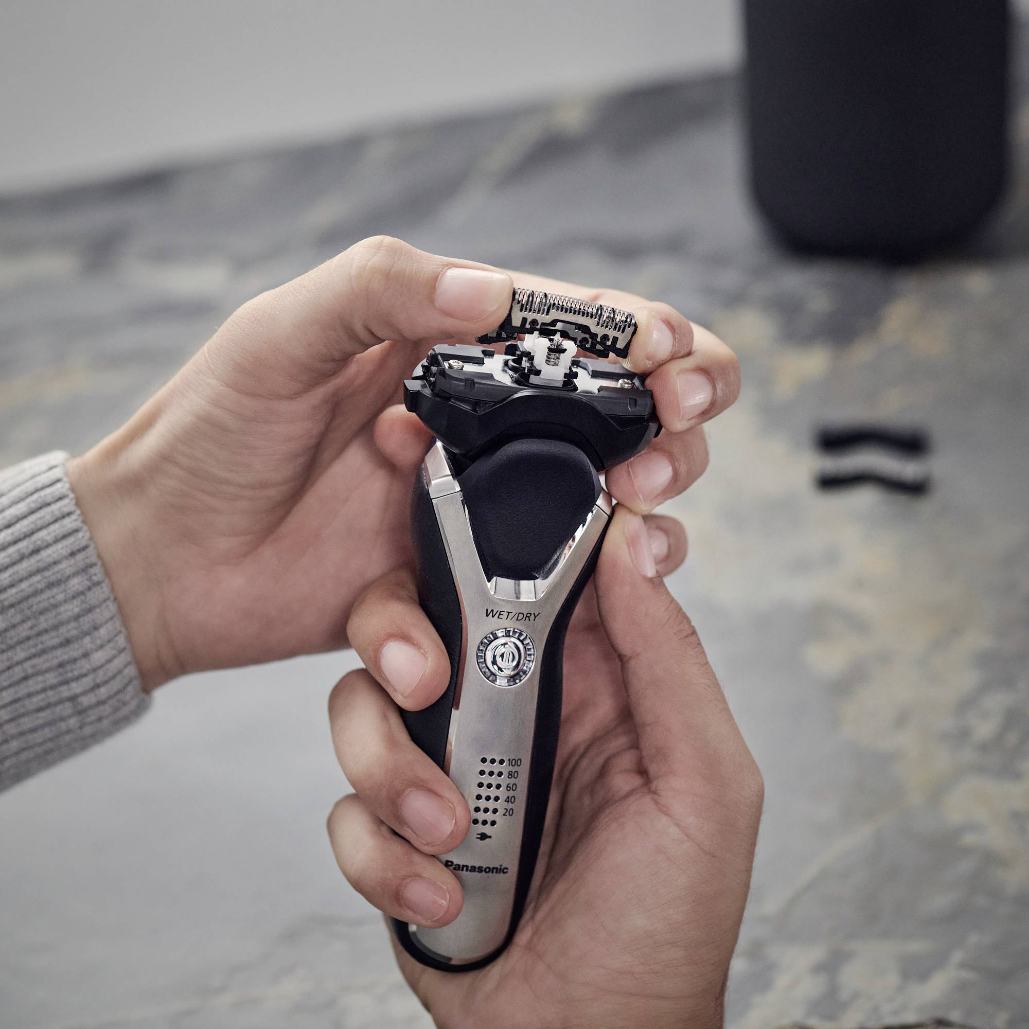A person is holding an electric razor and examining the shaving head. The razor is resting on a grey table.