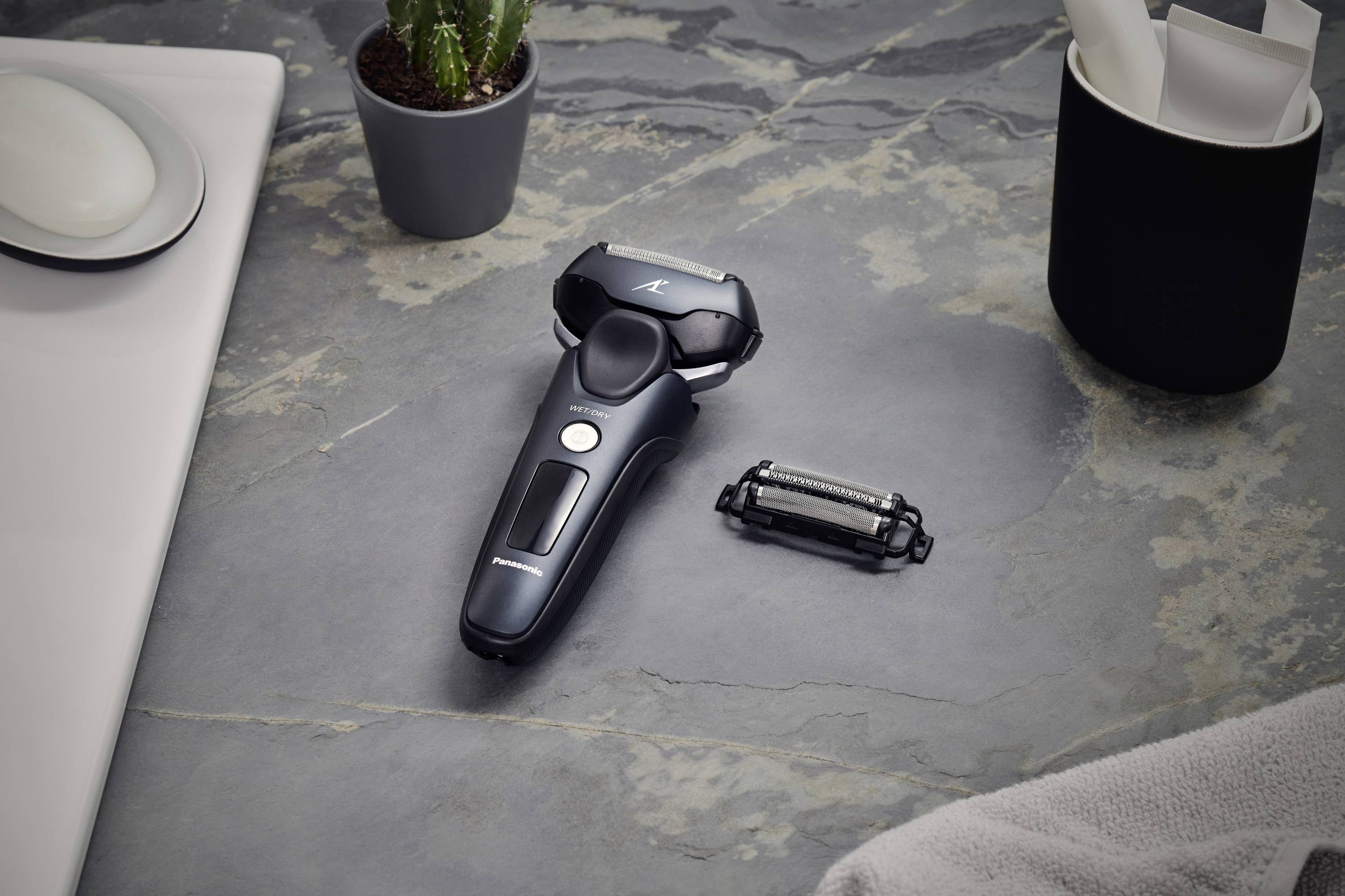 Electric shaver with additional cutting head on grey surface, positioned beside a plant and mugs, conveying a bathroom or grooming scenario.