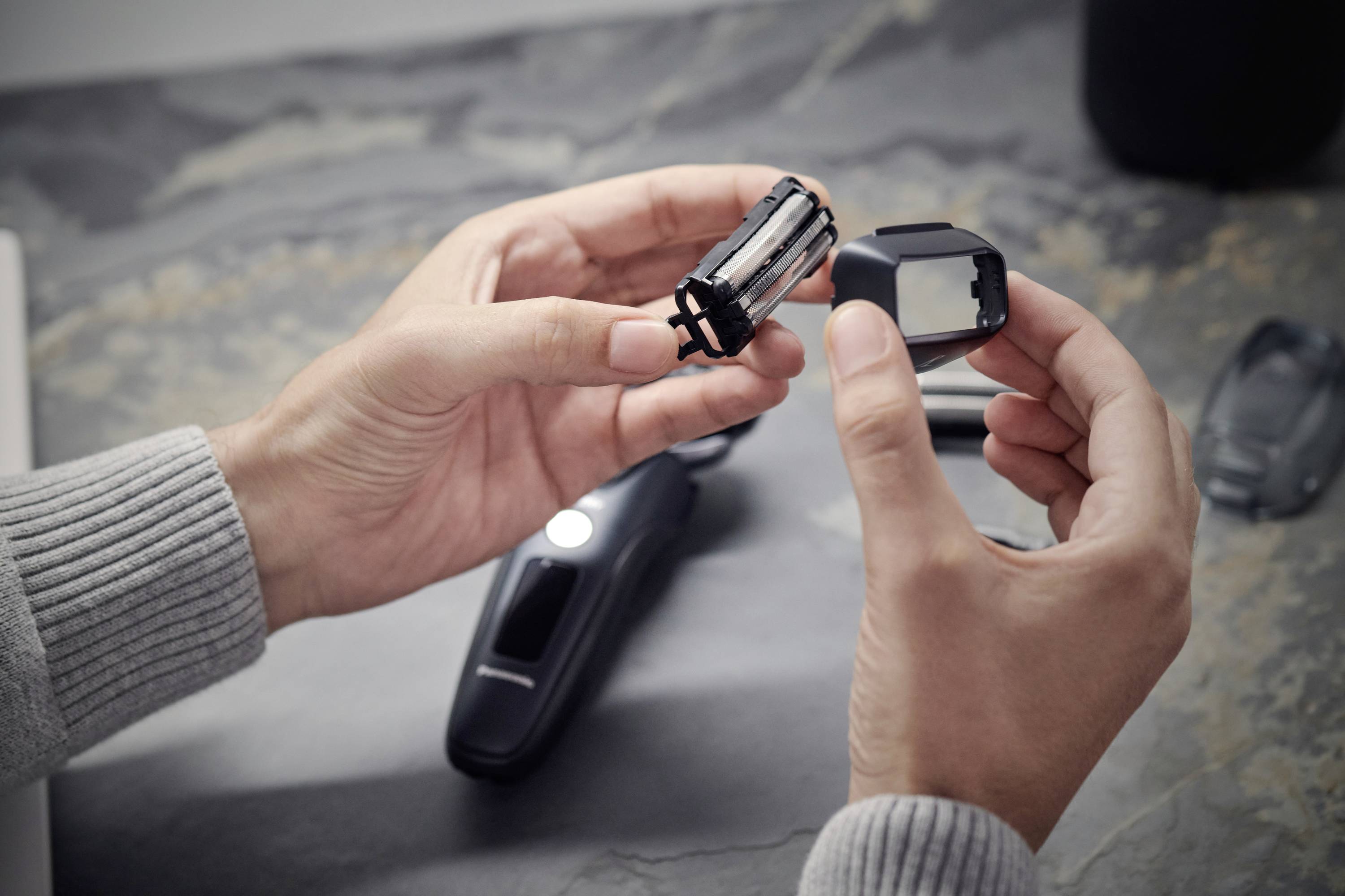 A person is holding spare parts of an electric razor in their hands, with the razor lying on the table in the background.