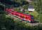 A red double-decker train is travelling through a green, wooded landscape beside a white house standing in the sunlight.