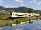 A yellow and white train travels along a lake in a rural landscape with hills and a blue sky in the background.