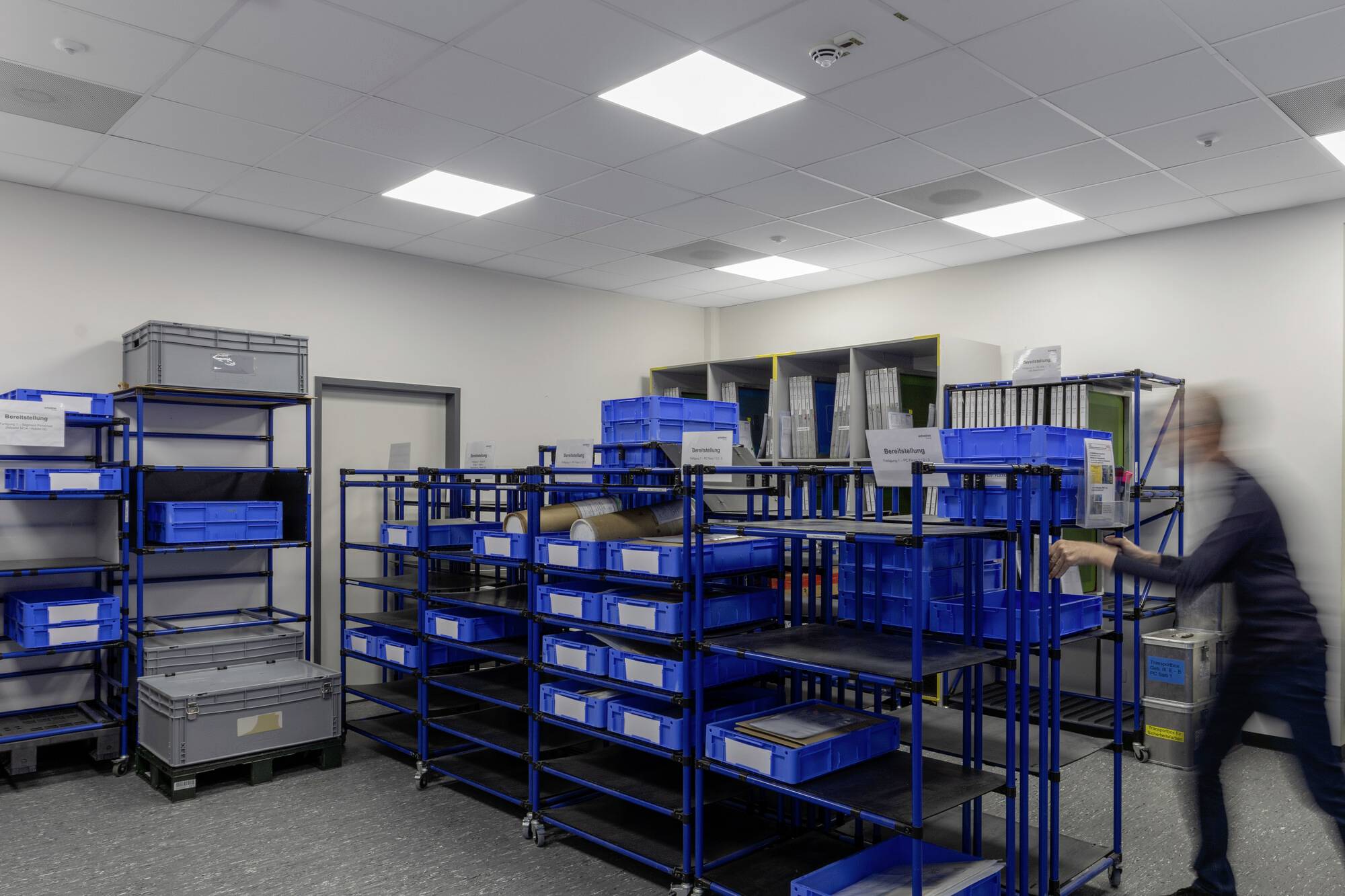 A blurry figure assembles shelves with blue plastic boxes in a storage room, surrounded by additional shelving and crates.