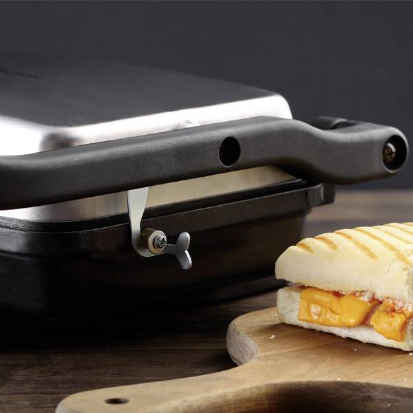A toasted sandwich with melted cheese is being pressed in a contact grill. It lies on a wooden board and is partially visible.