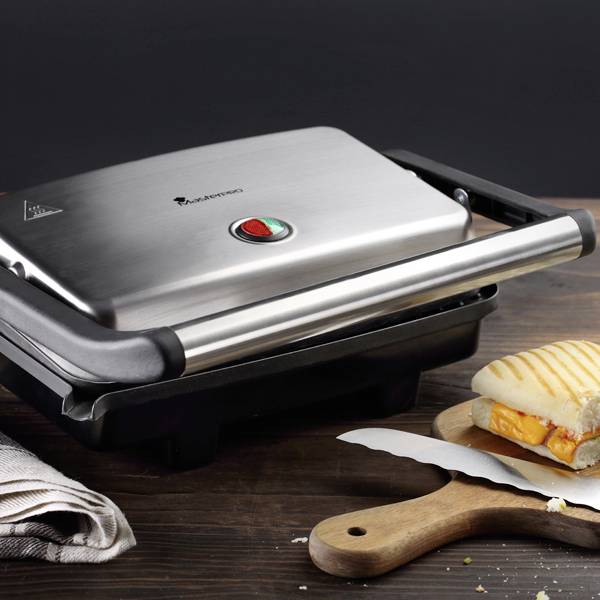 A sandwich maker on a wooden table displays a melted cheese sandwich on a wooden board. A tea towel lies beside it.