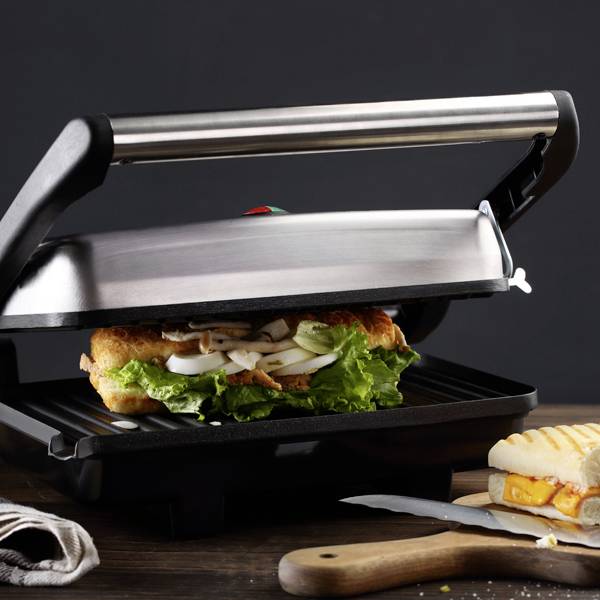 Sandwich toaster with a closed lid, toasting a sandwich with salad and mushrooms, with half a toasted cheese sandwich beside it.