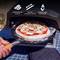 A person is sliding a pizza with mushrooms and mozzarella into a pizza oven, using a stainless steel pizza peel with an extra-long handle.