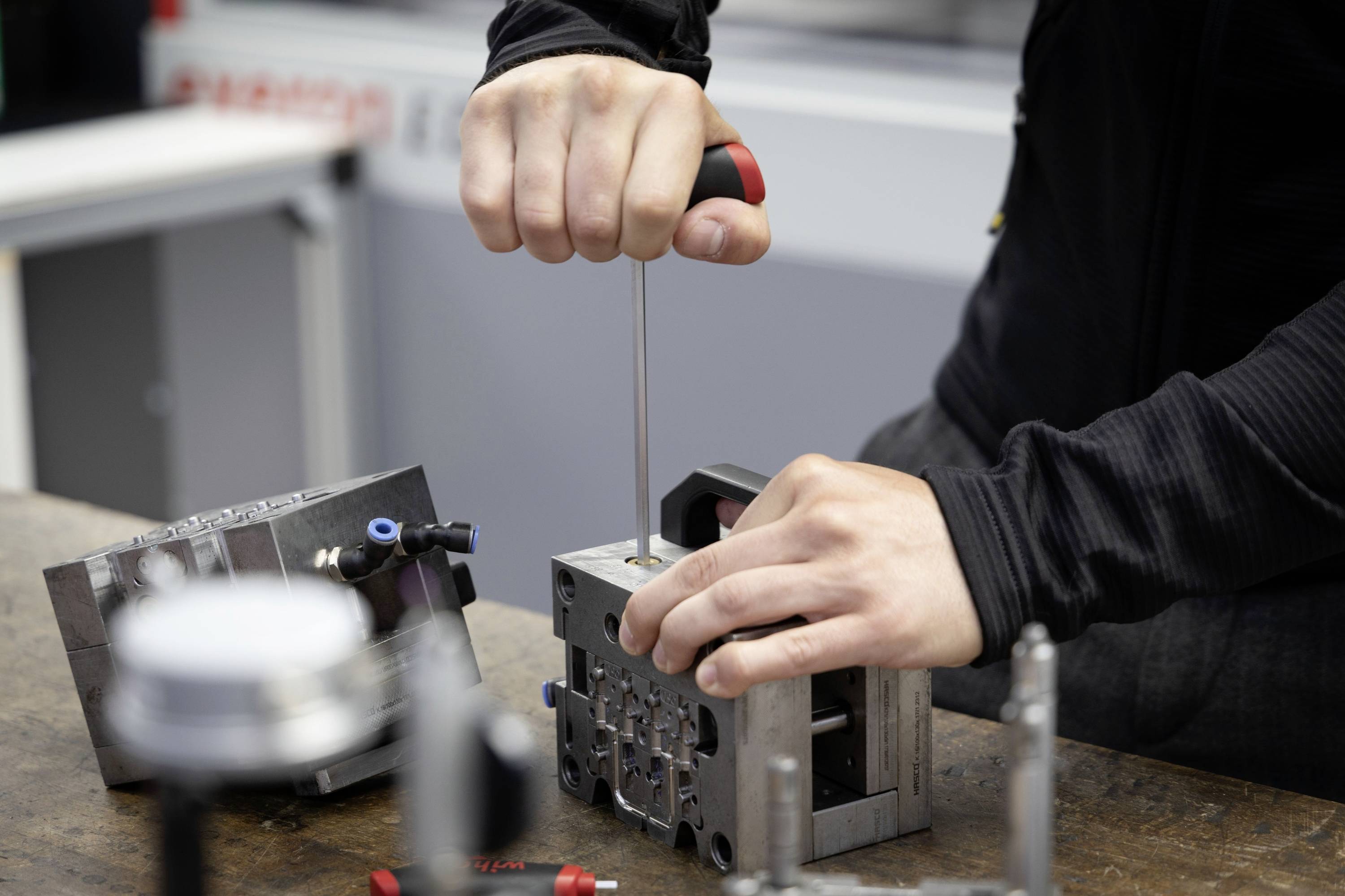 A person is aligning a technical component on a workbench using a screwdriver, surrounded by metal parts and tools.
