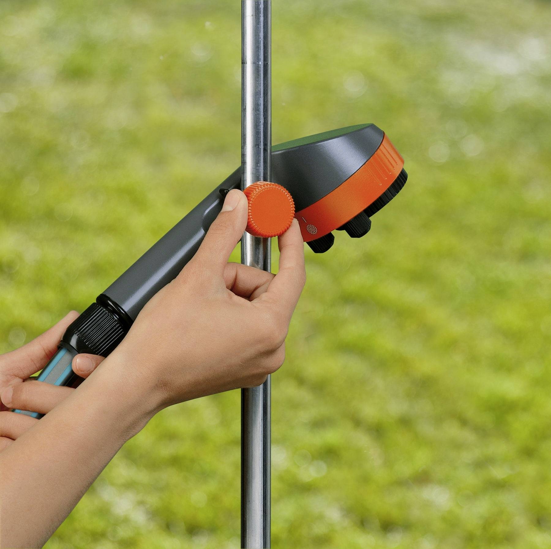 A person adjusts a clamp on a vertical metal pole outdoors, holding a tool with an orange dial and hose attachment, set against grass.