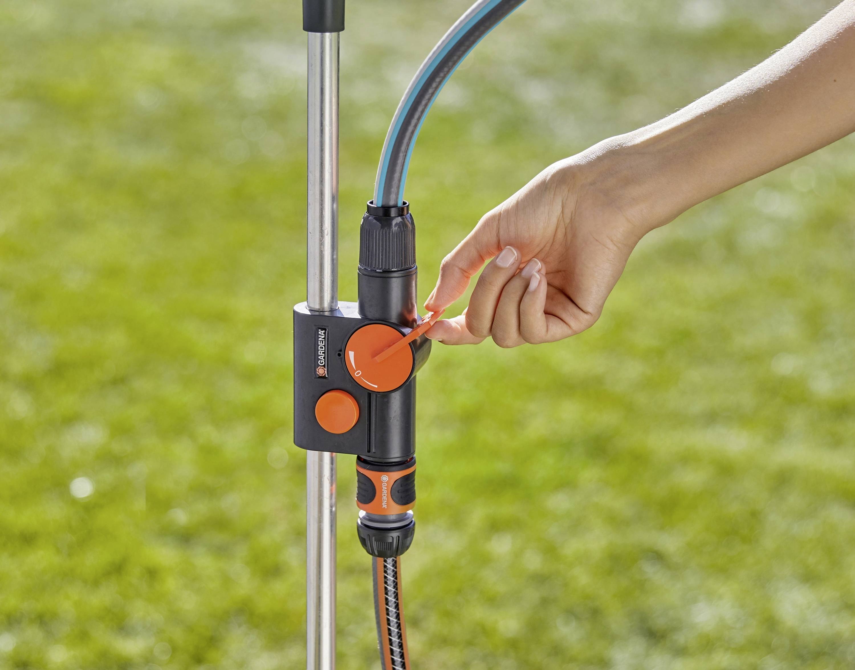 A hand adjusts a control valve on a garden water hose setup, with a green lawn in the background.