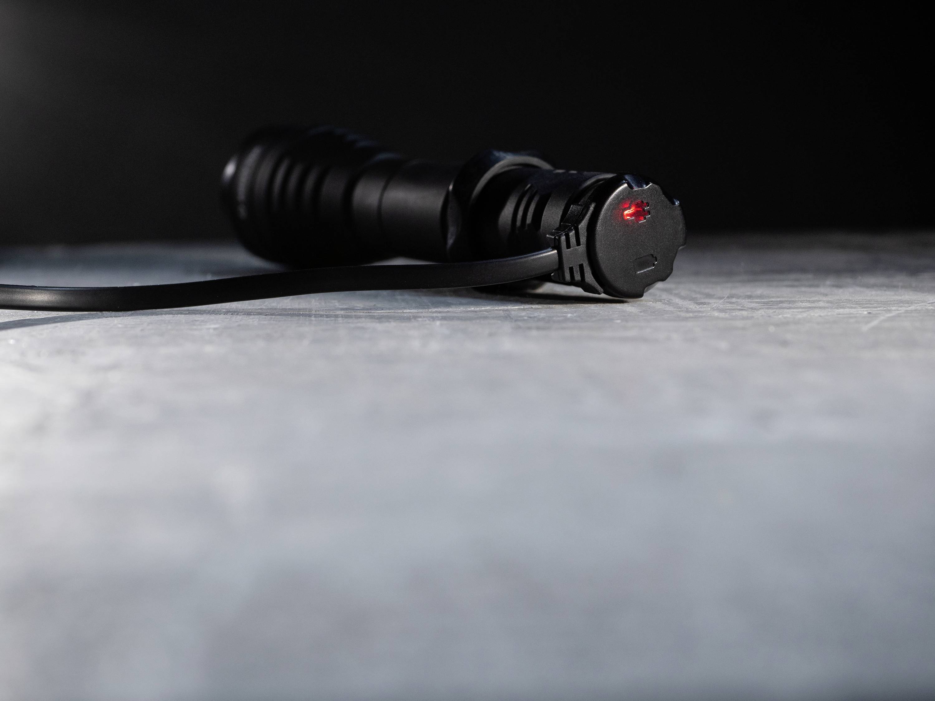 Black torch lies on a grey surface, showing a slightly glowing red light at one end, indicating low battery.