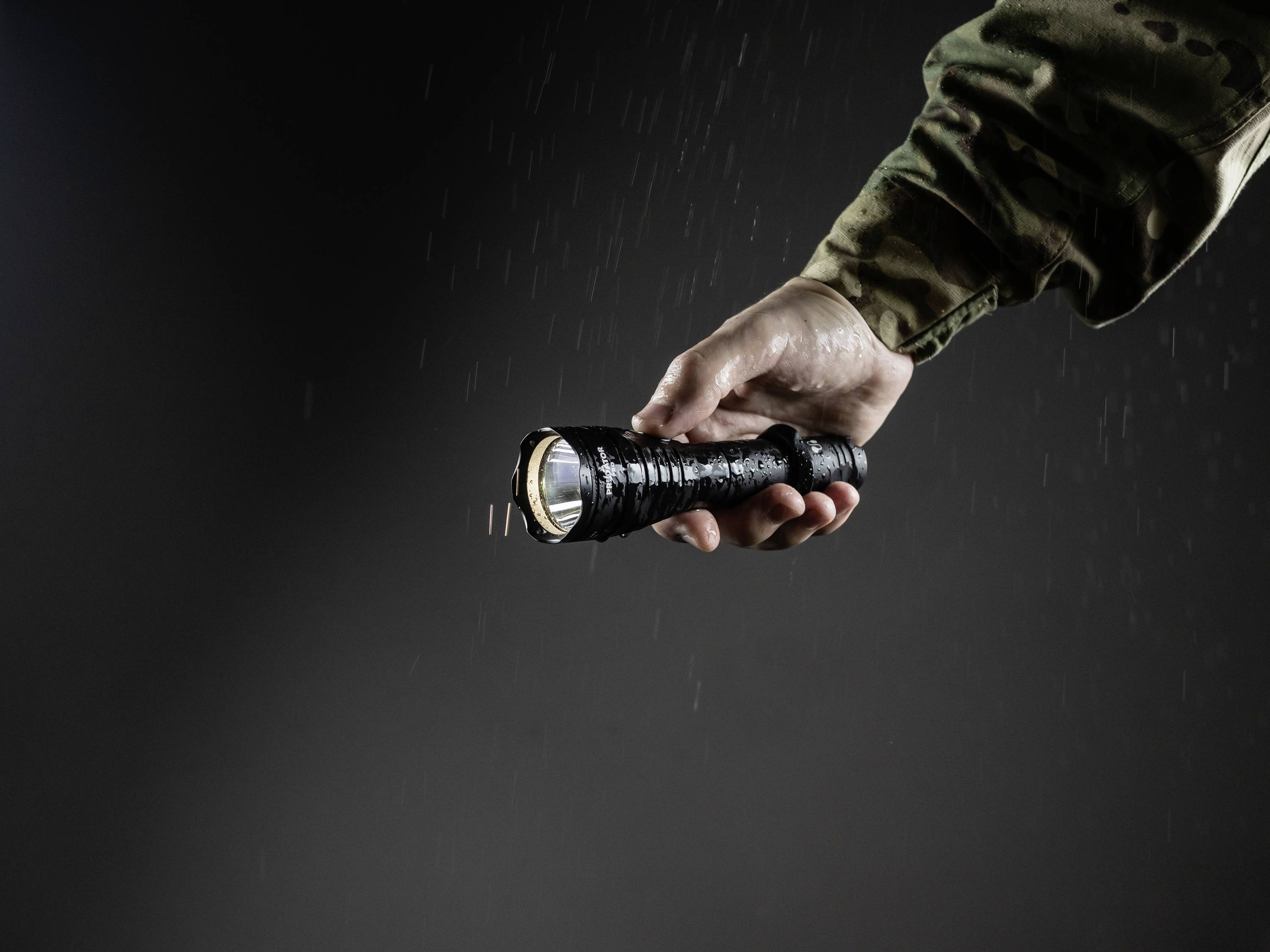 A hand in camouflage clothing holds a black torch, illuminating against a dark, rainy background.