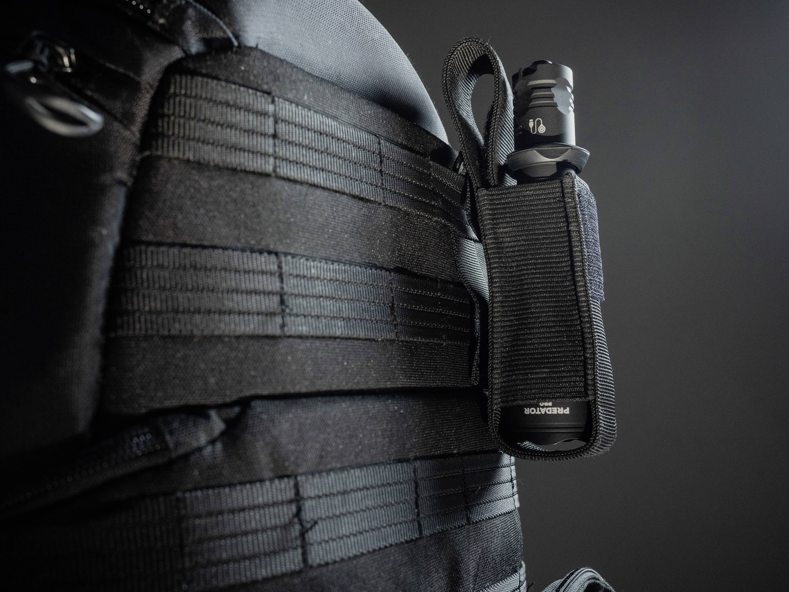 A black rucksack with MOLLE system. A torch is secured in a matching holder on the side. Background dark grey.