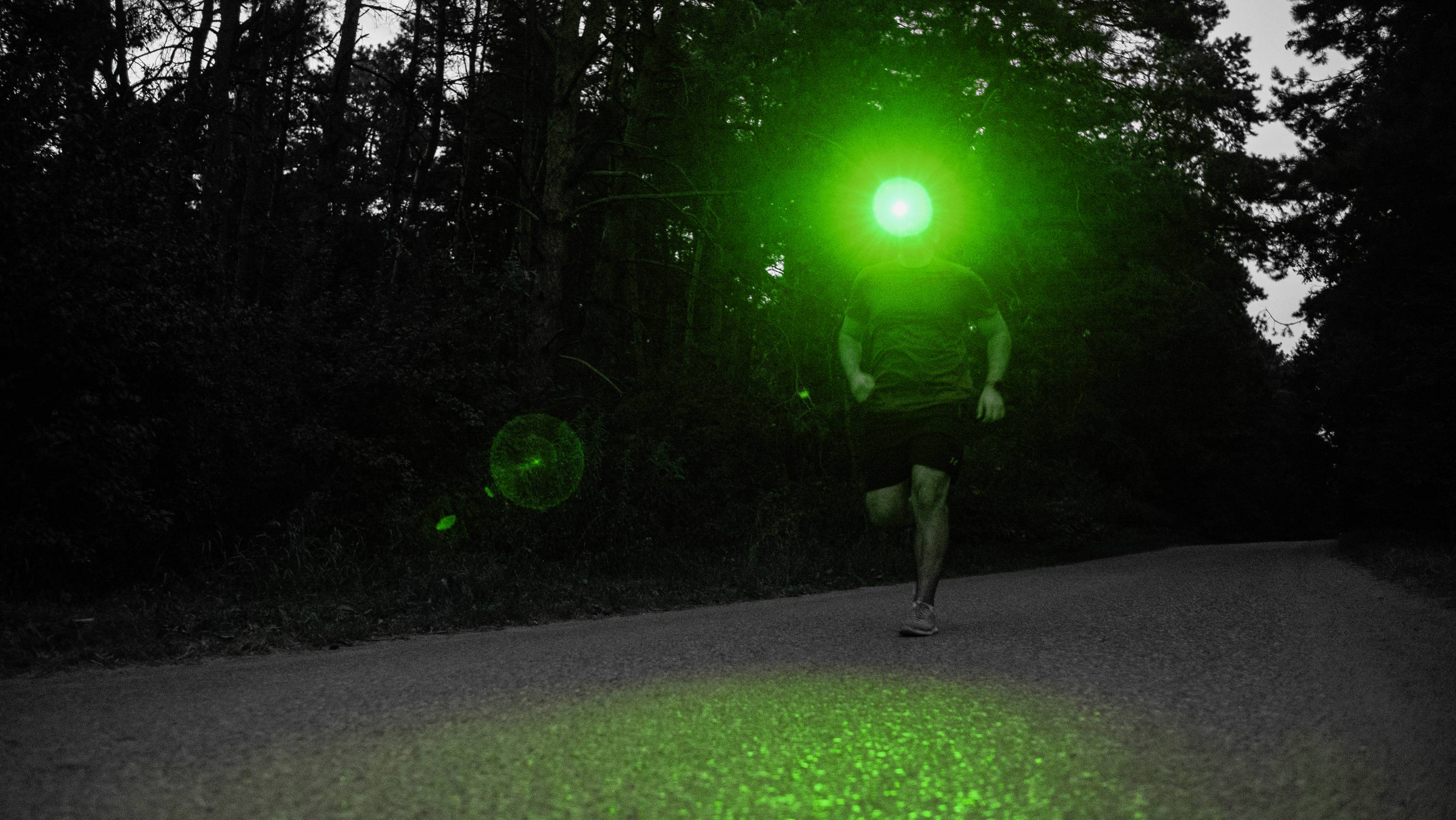 A person is jogging at night on a road through a woodland. A green light shines in front of their face, making them visible in the darkness.