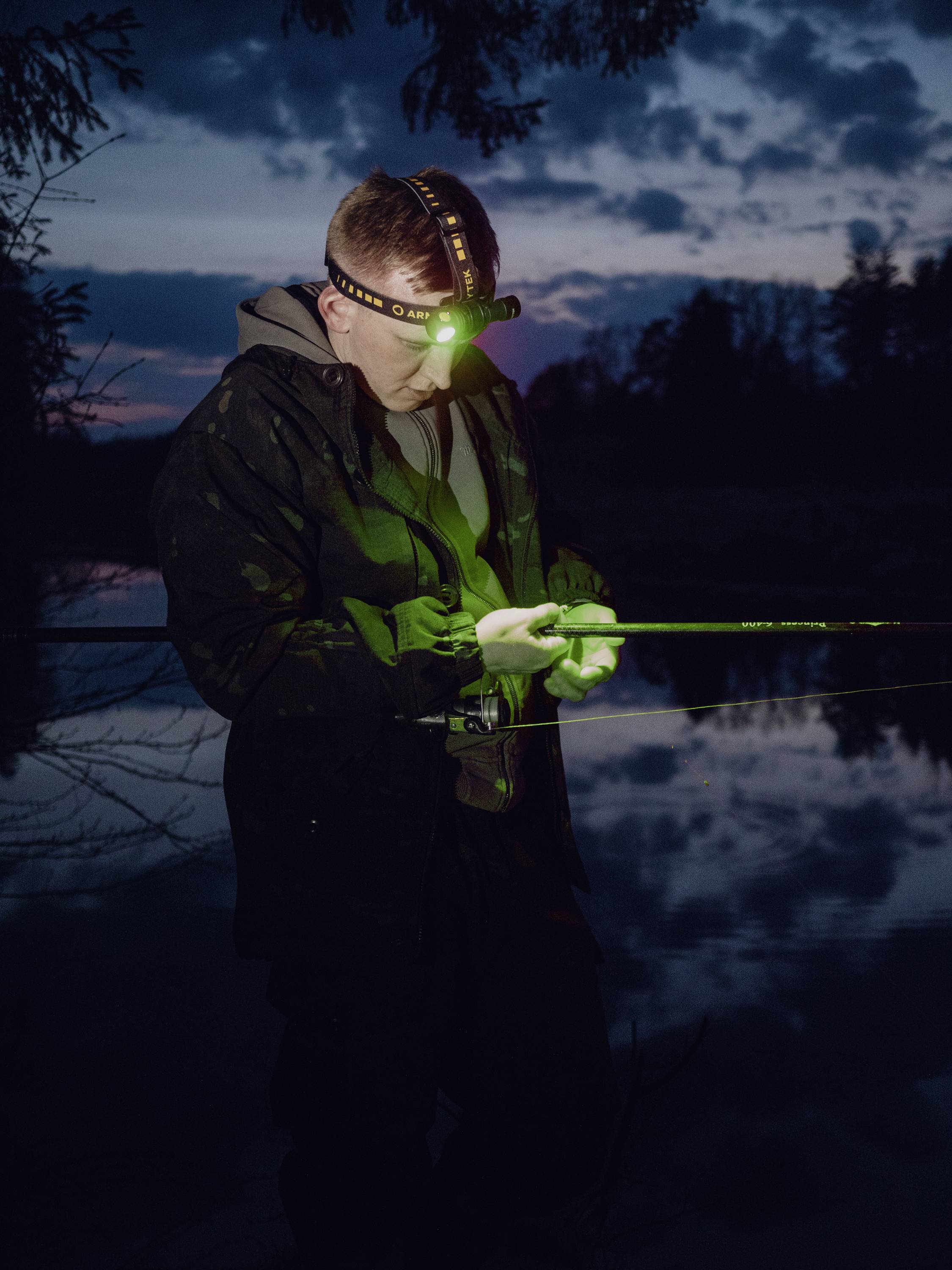 A person is fishing at night by a lake. They are wearing a head torch illuminating their fishing equipment. The sky is dark in the background.
