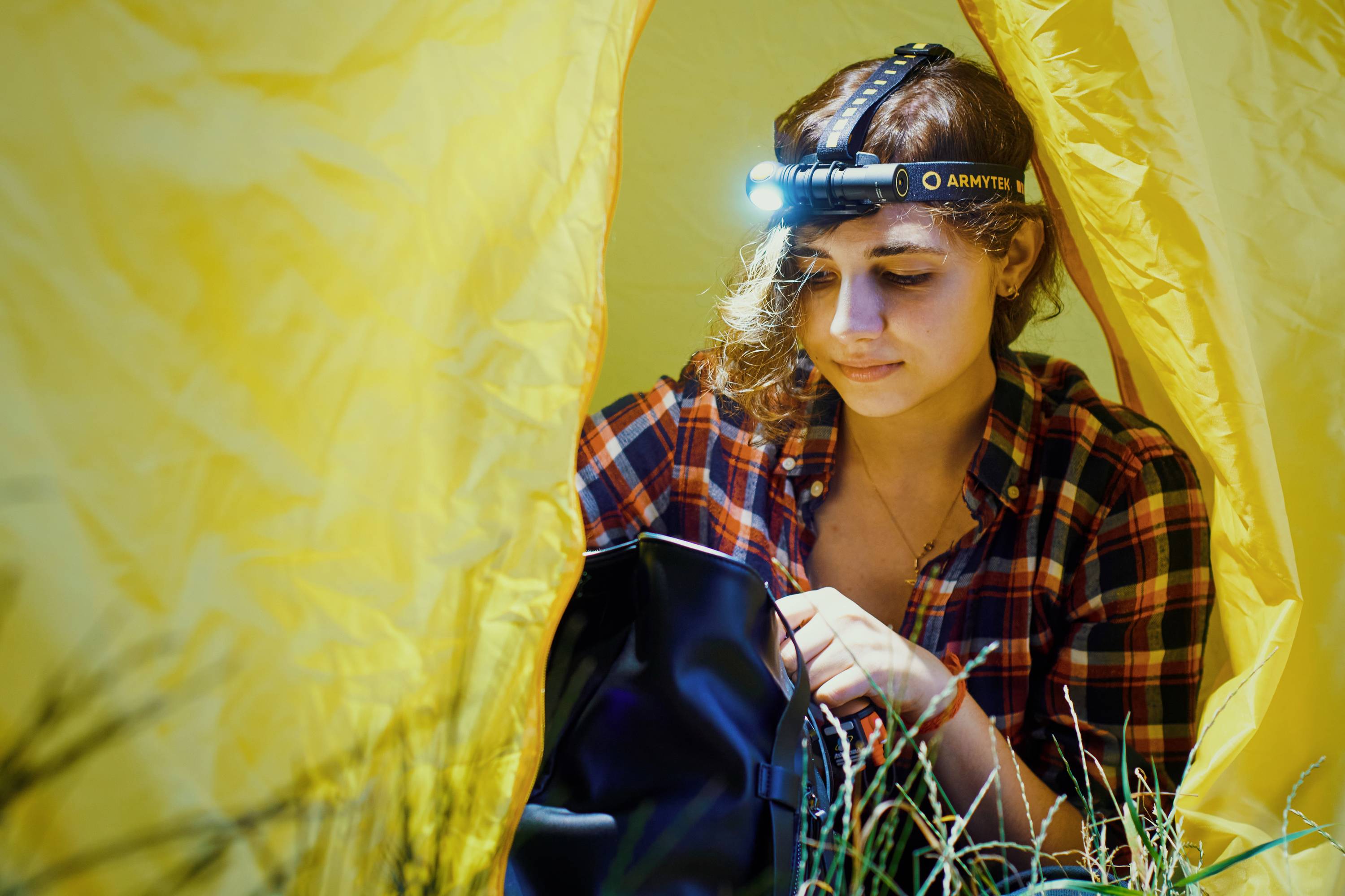 A person in a checked shirt sits inside a yellow tent, shining a head torch into a bag. Grass can be seen outside.