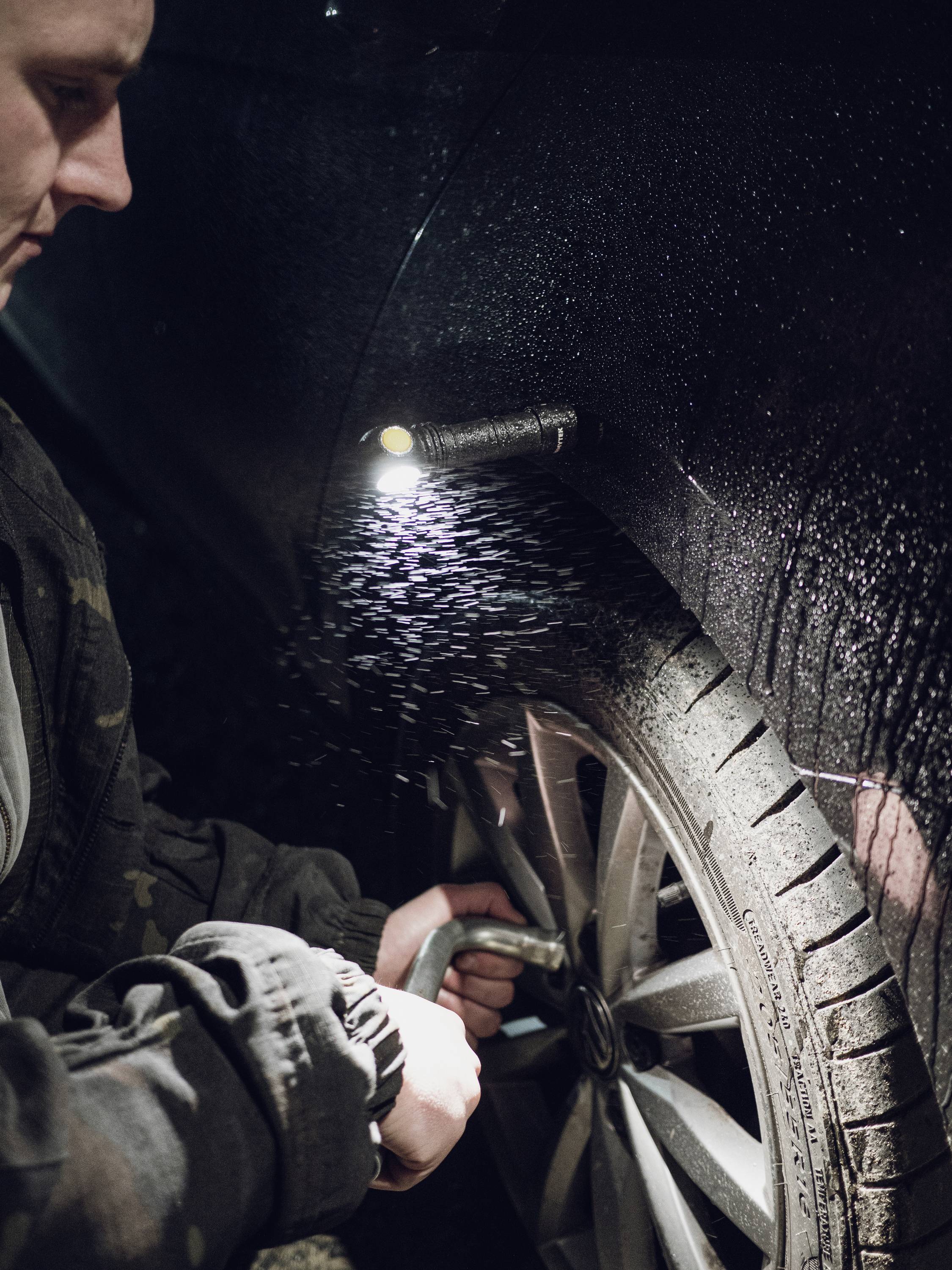 A person is inflating a car tyre at night, illuminated by a small lamp. Raindrops glisten on the bodywork.