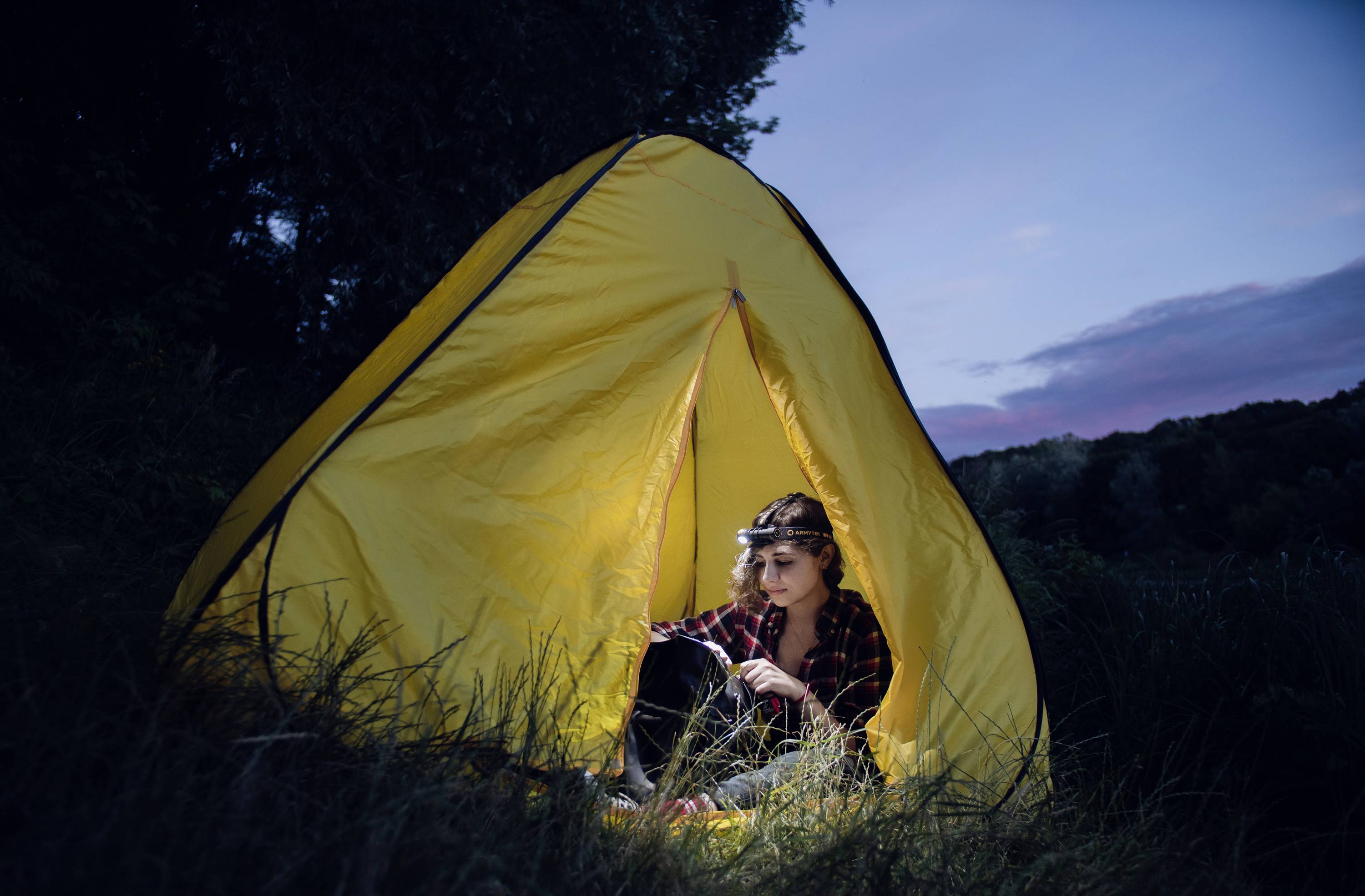 A person sits in a yellow tent on the grass at dusk, wearing a head torch and gazing thoughtfully into the distance.
