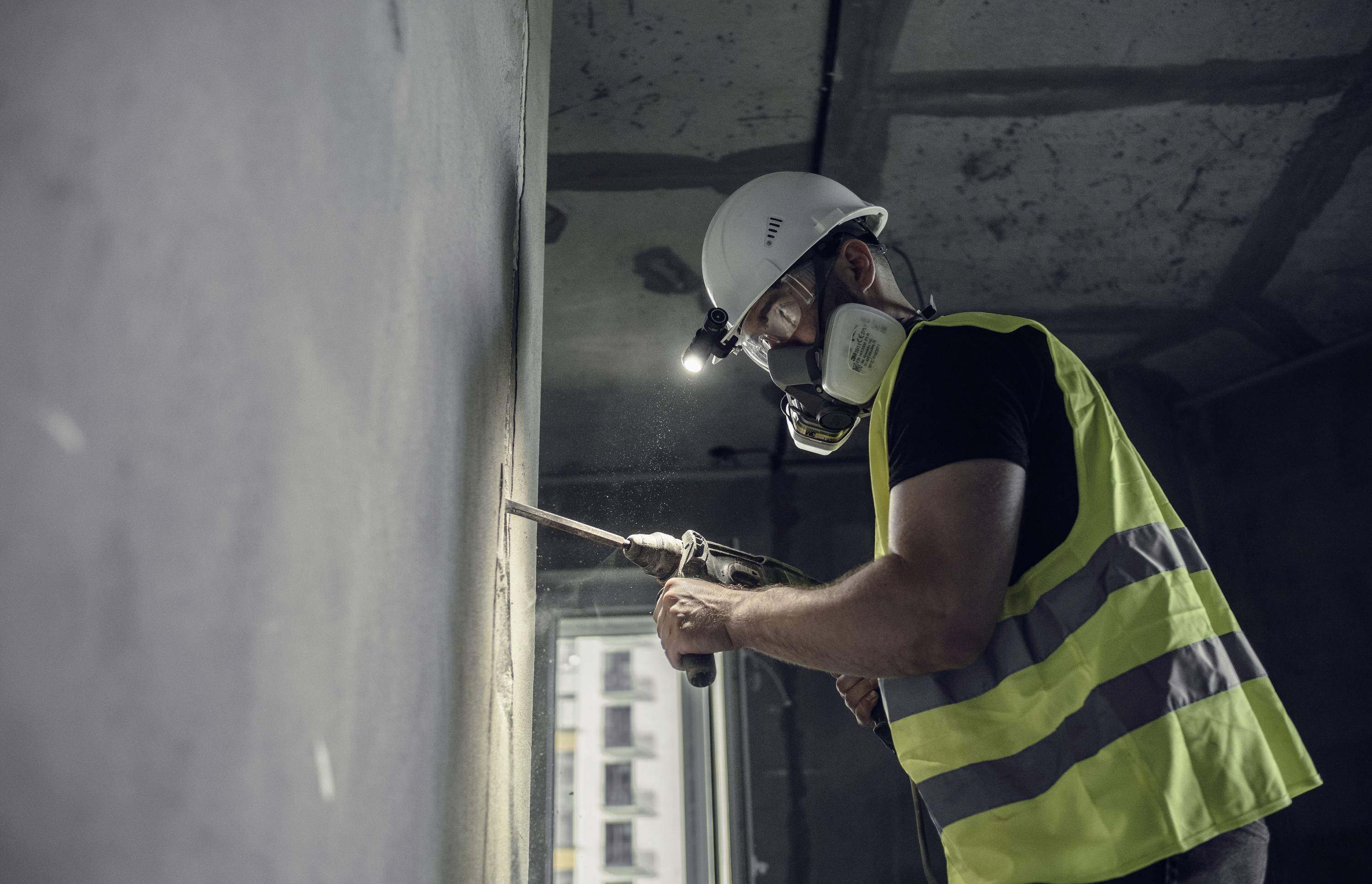 A construction worker wearing a protective mask and high-visibility vest is carrying out drilling work on a concrete wall in an unfinished building.