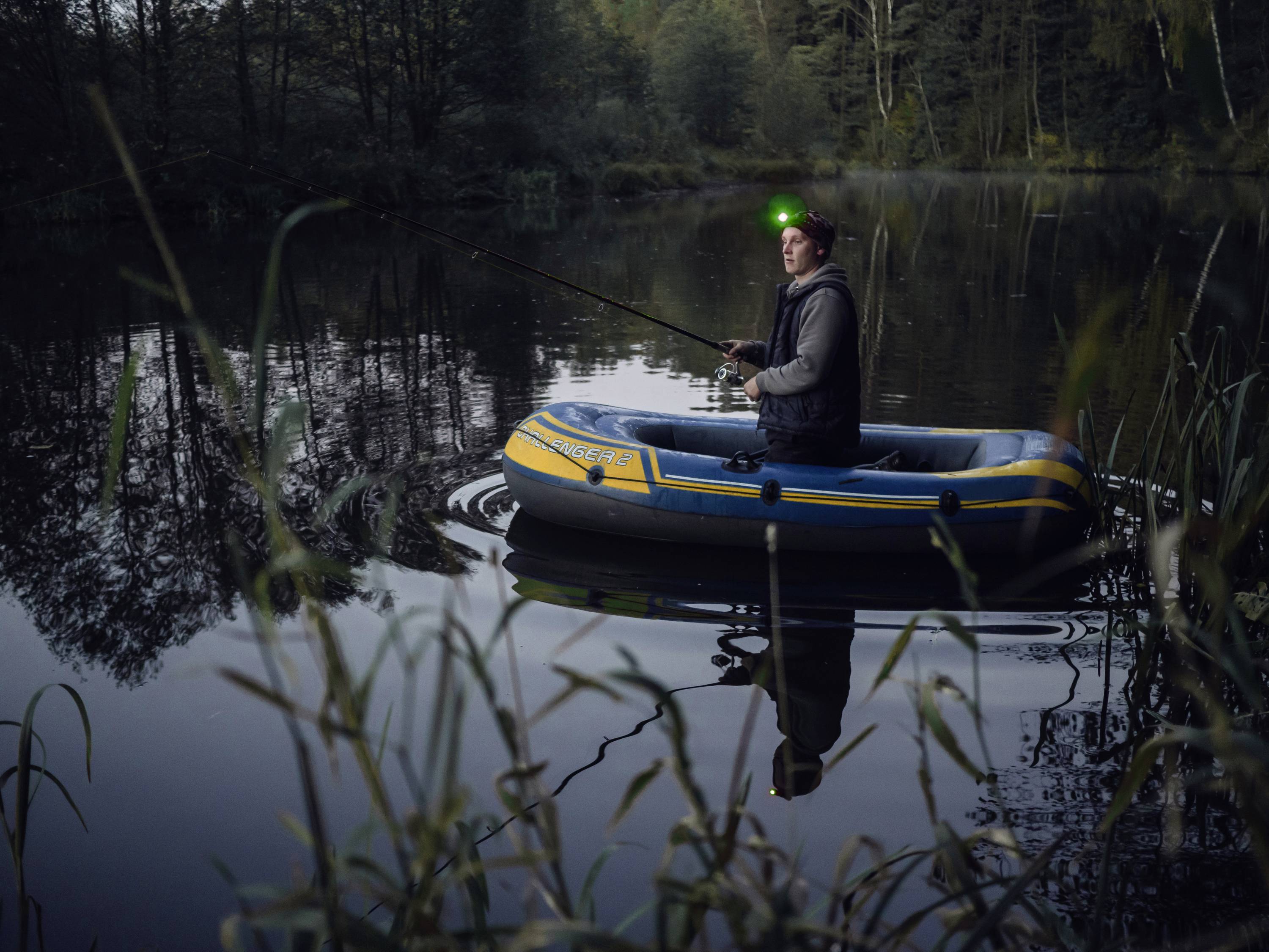 A person is fishing at night on a calm lake in an inflatable dinghy, wearing a head torch. Trees are reflected in the water.
